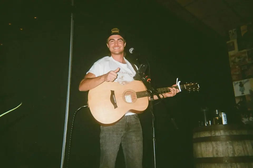 Film photo of Nik Poli grinning with a thumbs up at the mic, acoustic guitar, on a small bar stage