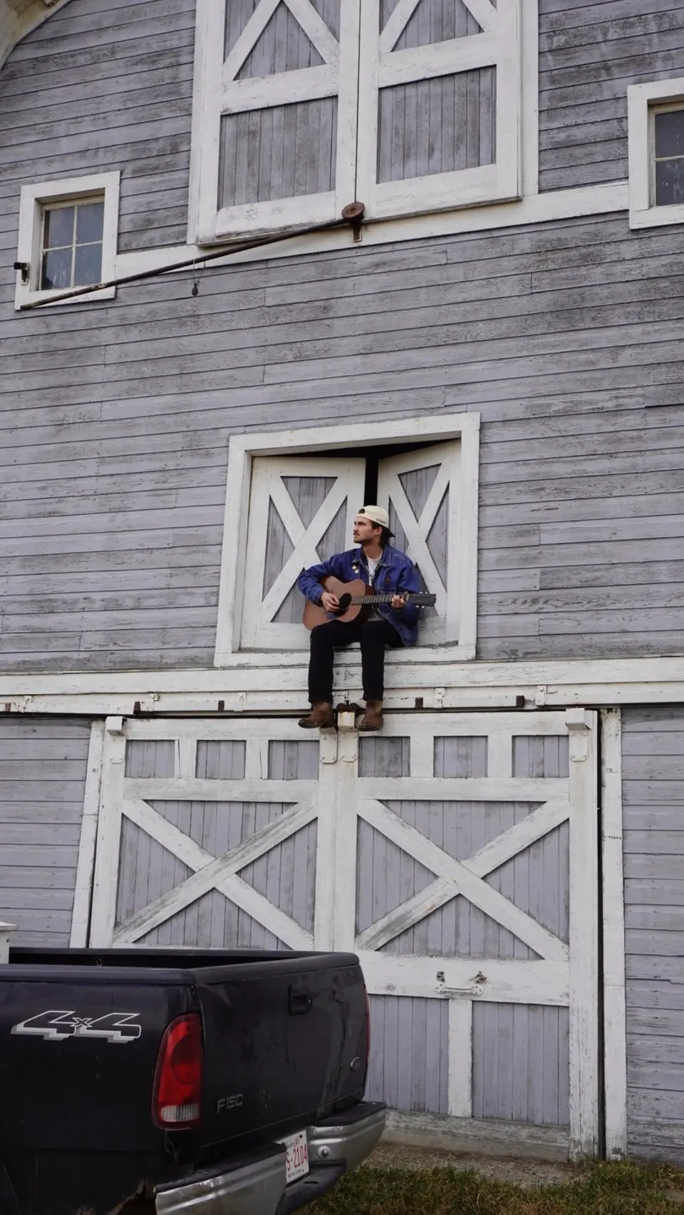 Nik Poli sitting in the hayloft door of a weathered grey barn, pickup truck below