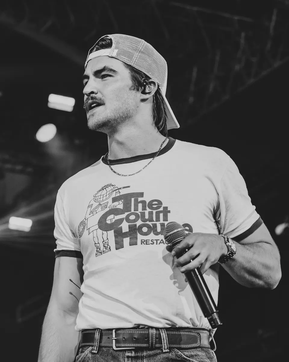 Black and white portrait of Nik Poli holding the mic, backwards cap, Cowboys 2026
