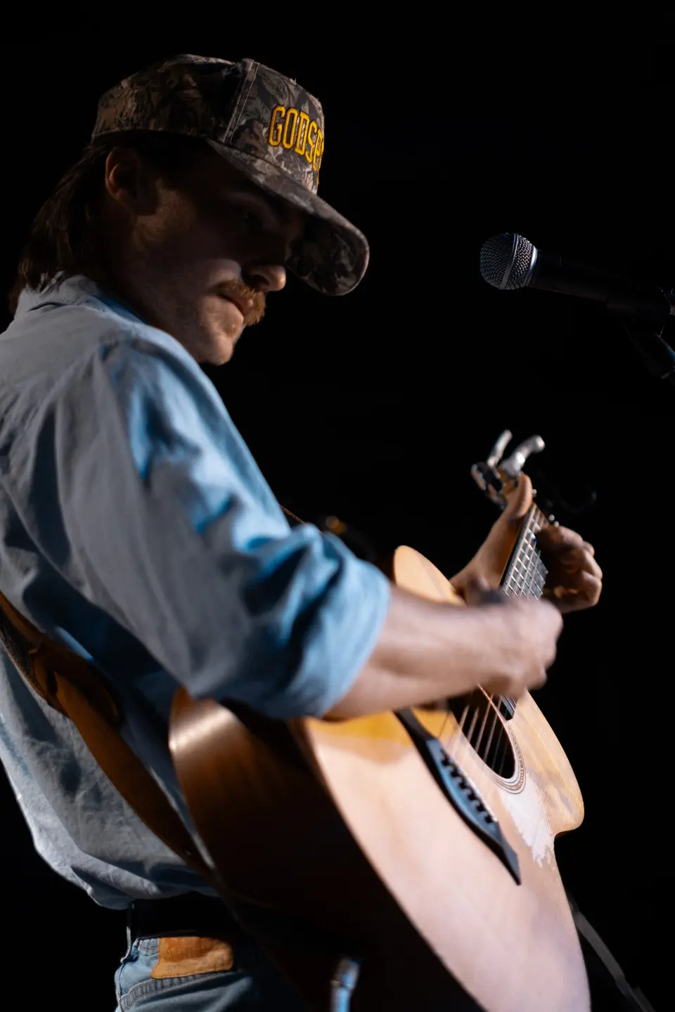 Nik Poli in a blue shirt and cap, head down, playing acoustic guitar at the mic