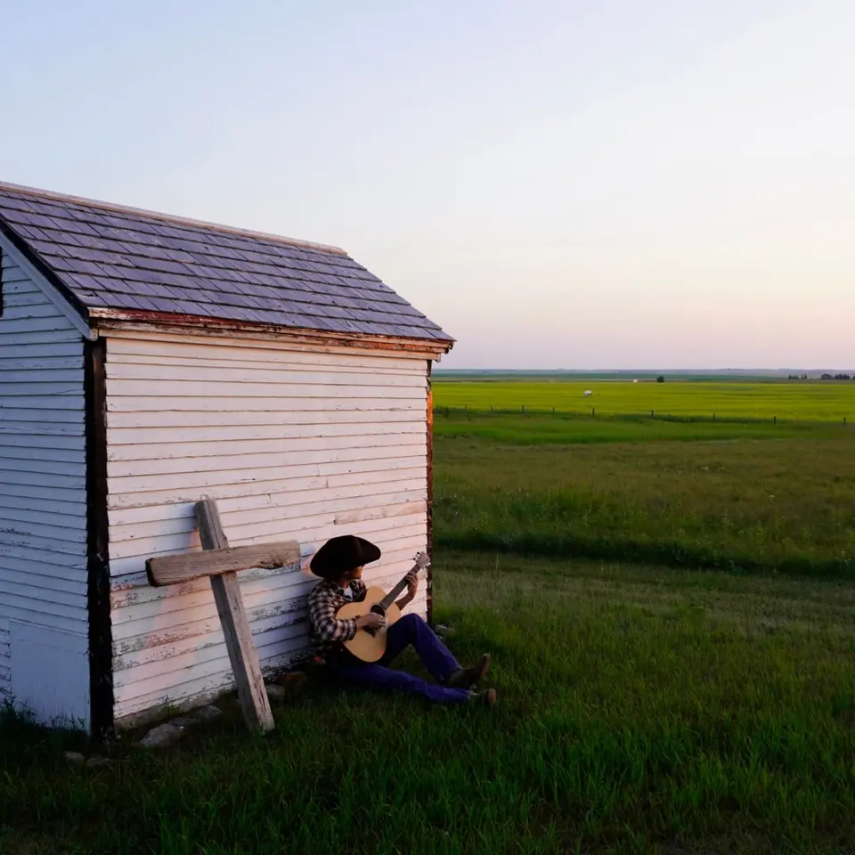 Nik Poli playing guitar beside a white shed on the prairie at dusk