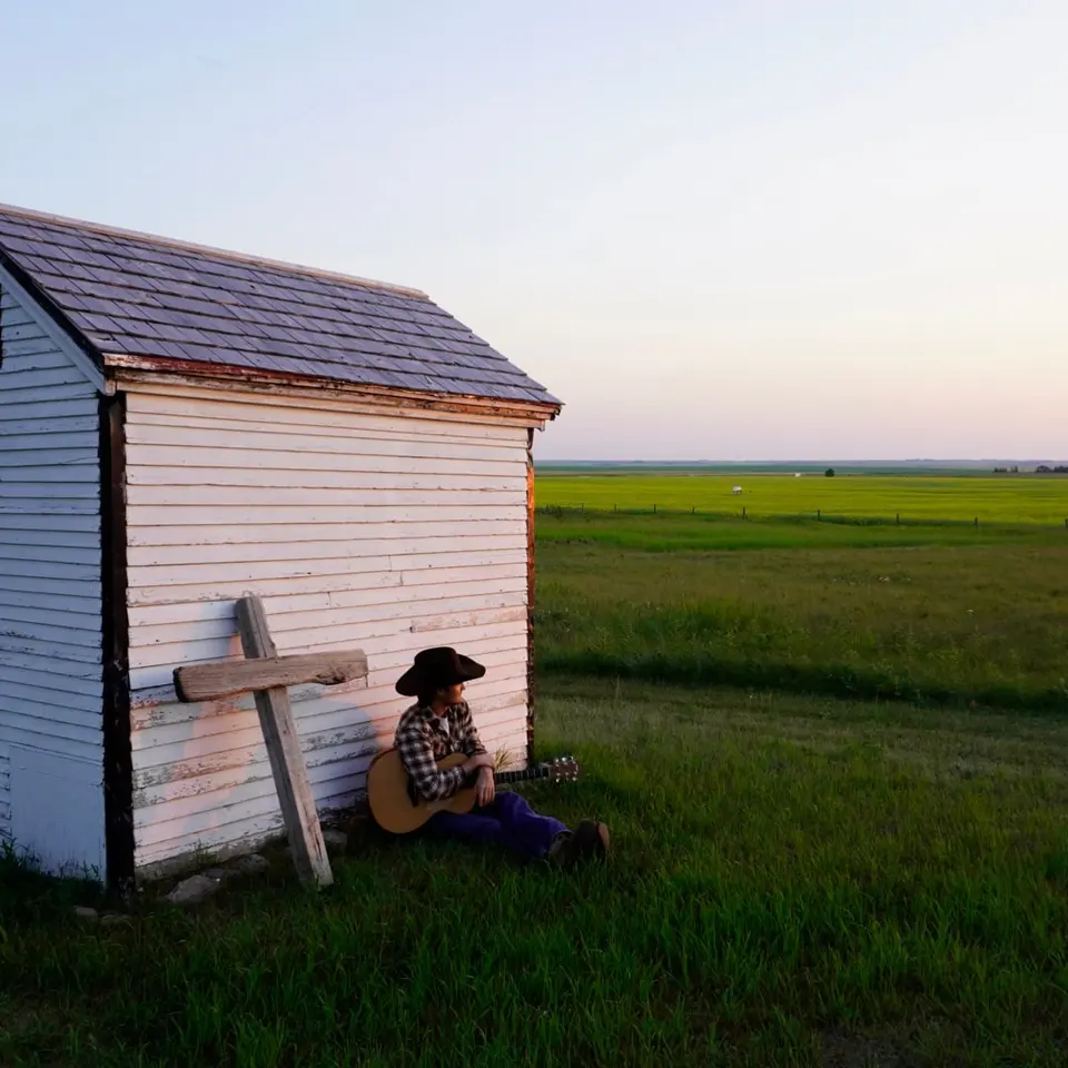 Nik Poli sitting against a white prairie shed at sunset, wooden cross beside him