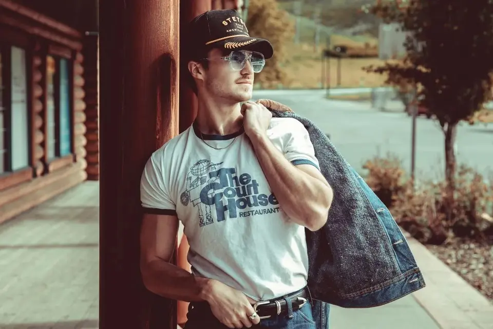Nik Poli leaning on a log-cabin post in a Stetson cap and sunglasses, denim jacket over one shoulder