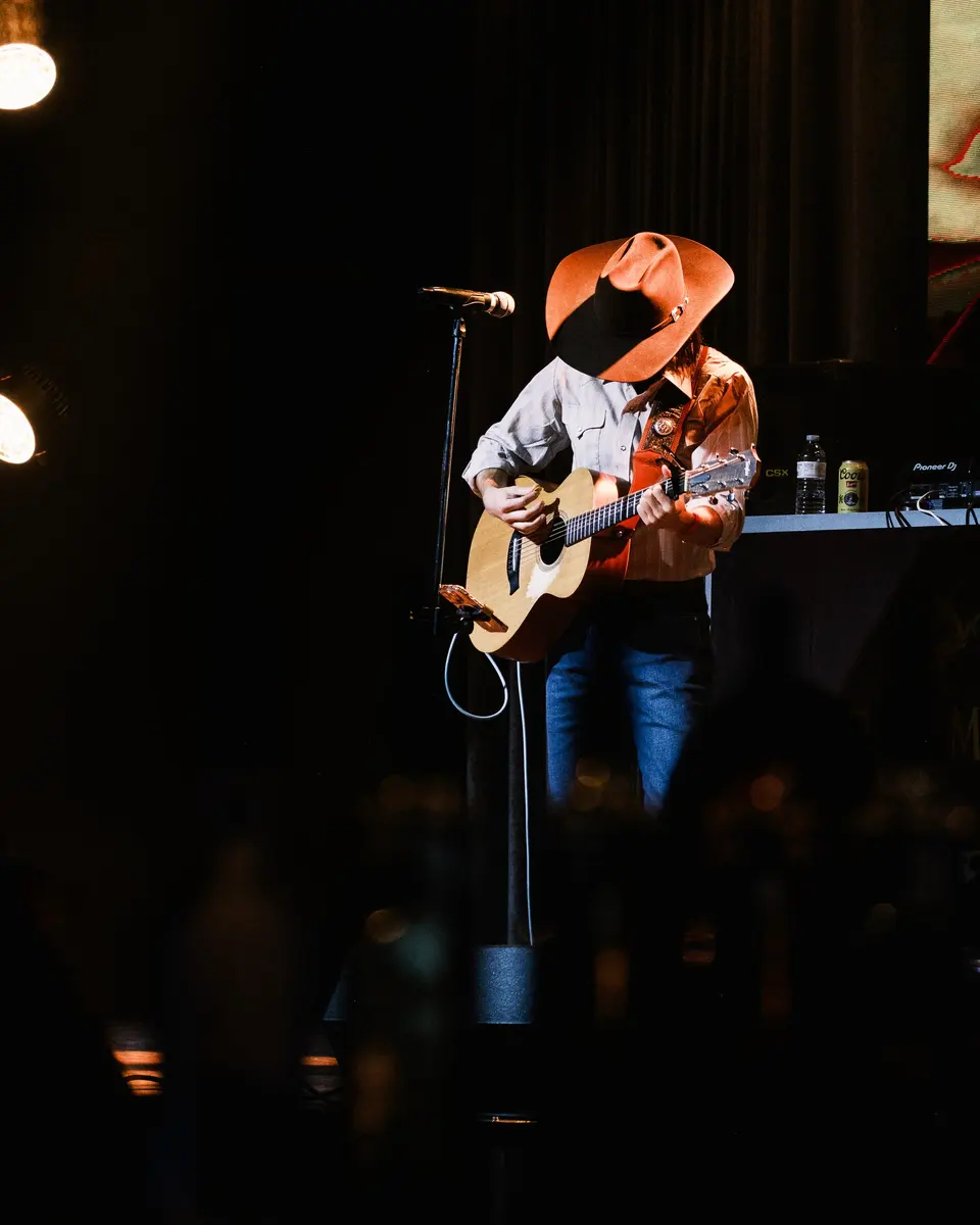 Nik Poli in a cowboy hat on a dark stage, acoustic guitar, single spotlight