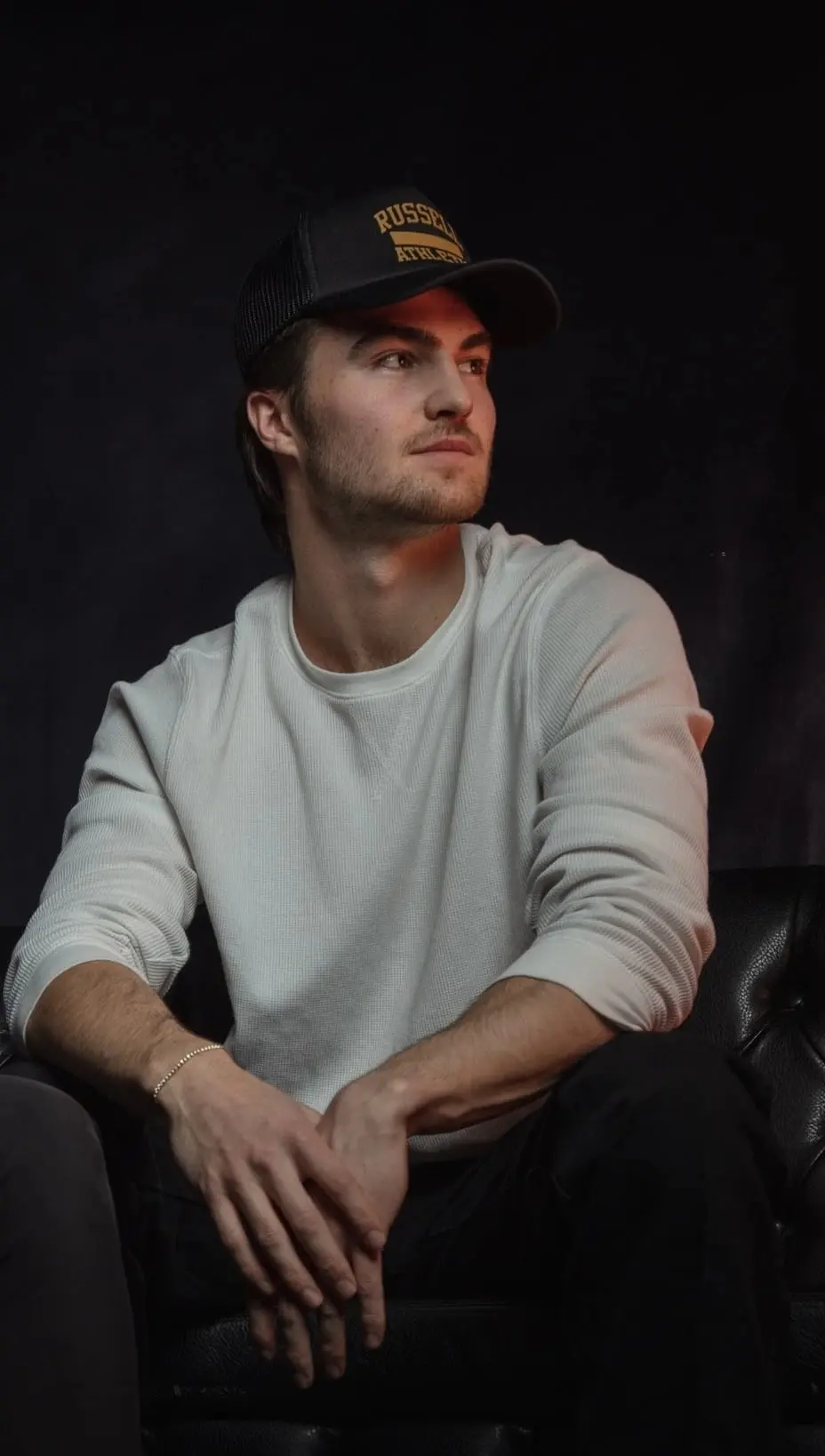Studio portrait of Nik Poli seated on a black leather couch in a trucker cap and white sweater, looking off to the side